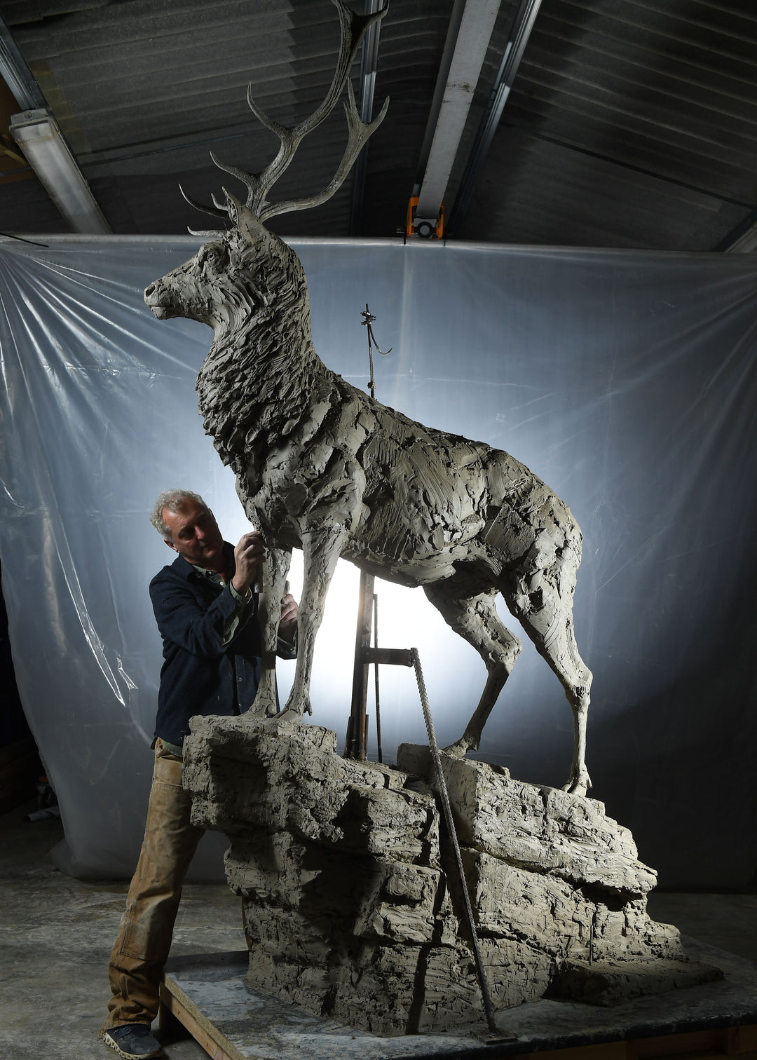 ‘Red Stag: Life Size’ by Hamish Mackie, a monumental limited-edition bronze wildlife sculpture depicting a mature Scottish Highland red deer stag standing alert on a rugged rocky outcrop. Featuring an imposing set of branching antlers, a powerful muscular body and richly textured coat, the sculpture captures the strength, dignity and commanding presence of a stag surveying its territory. Photographs show the original hand-modelled clay sculpture prior to bronze casting.