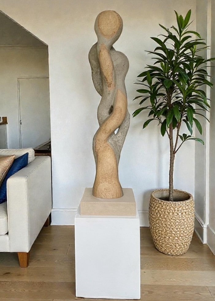 Carved abstract figurative sculpture formed from a single block of Blue and Buff Ancaster Weatherbed stone, with intertwined vertical forms rising from an Ancaster stone plinth, featuring smooth flowing contours, subtle surface texture and balanced proportions designed for indoor display.