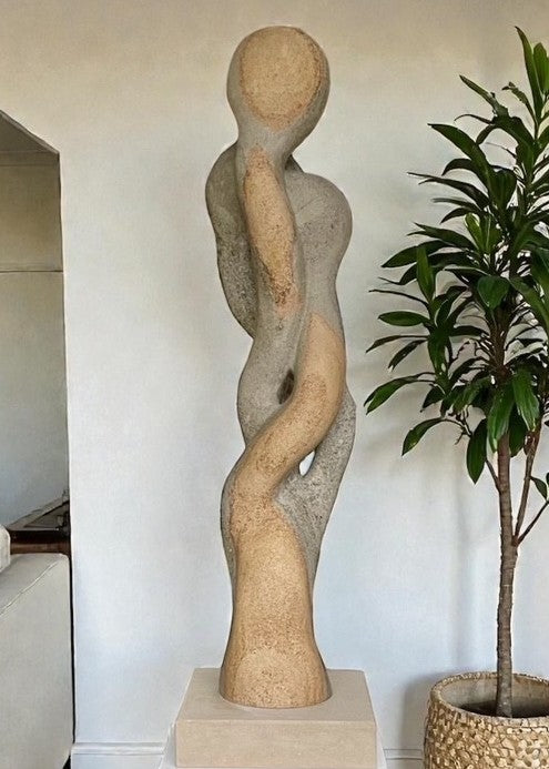 Carved abstract figurative sculpture formed from a single block of Blue and Buff Ancaster Weatherbed stone, with intertwined vertical forms rising from an Ancaster stone plinth, featuring smooth flowing contours, subtle surface texture and balanced proportions designed for indoor display.