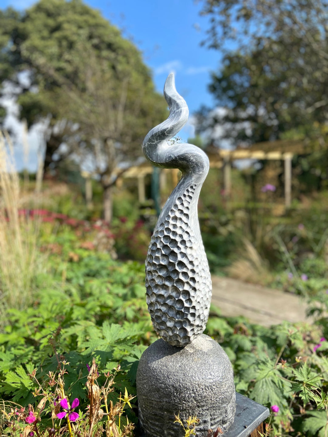 Release by Brendan Cullen, a hand-carved grey marble sculpture with a honeycomb-textured base that rises into a twisting, fluid spiral form, mounted on a smooth stone plinth, designed for outdoor display.