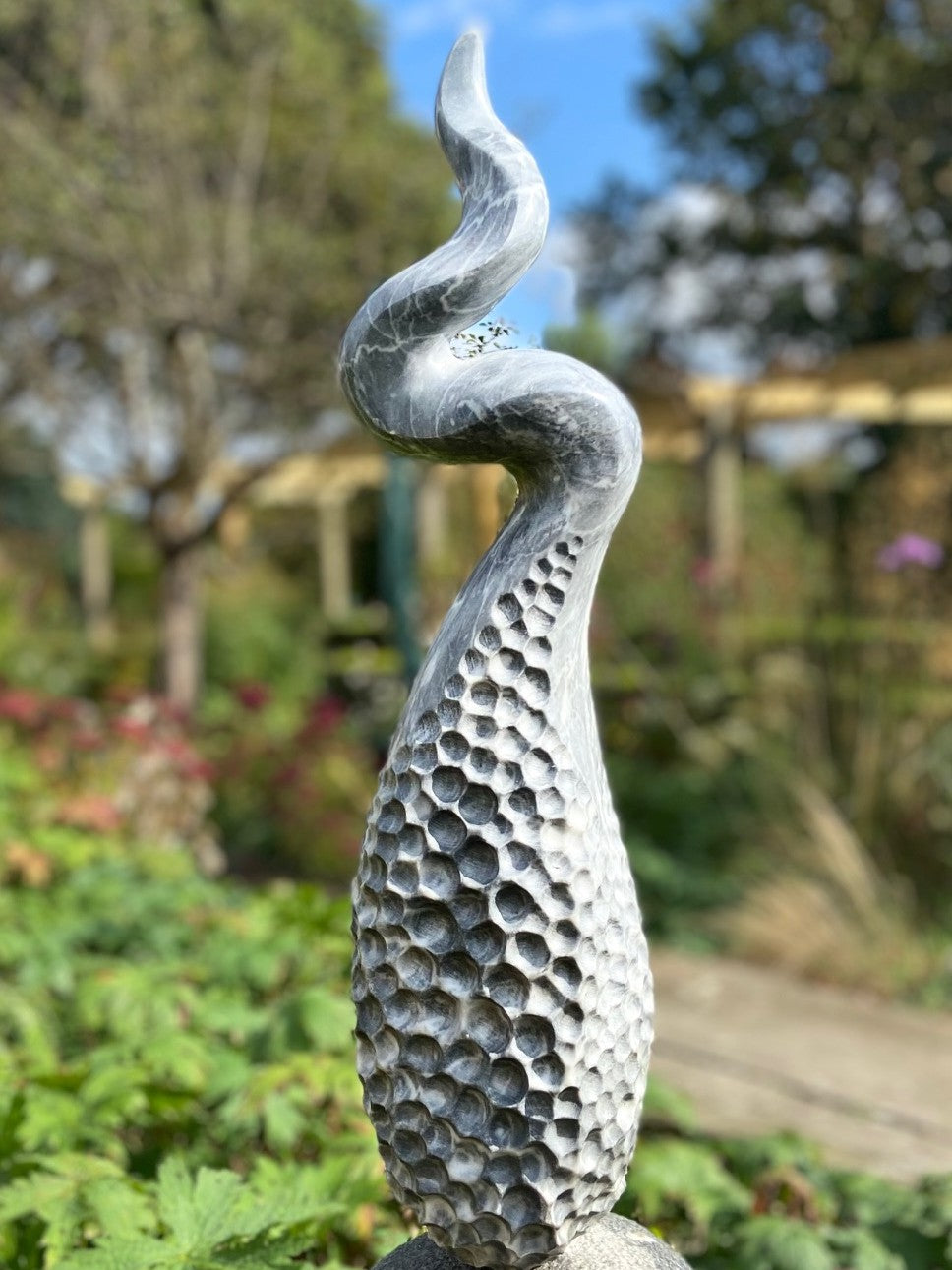 Release by Brendan Cullen, a hand-carved grey marble sculpture with a honeycomb-textured base that rises into a twisting, fluid spiral form, mounted on a smooth stone plinth, designed for outdoor display.