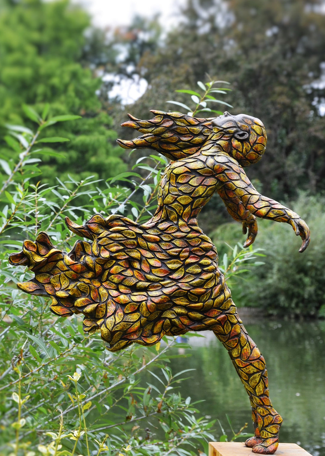 Jonathan Hateley Releasing bronze sculpture depicting a stylised female figure in motion, her form composed of layered leaf-like textures flowing outward as if caught in an autumn breeze. The figure leans forward with arms extended and head lifted, conveying a sense of release, renewal and movement, with warm bronze tones reflecting light across the textured surface against a natural outdoor setting.
