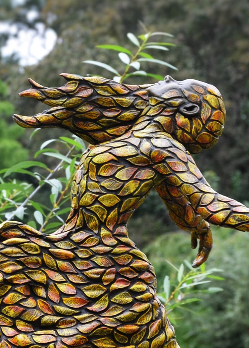 Jonathan Hateley Releasing bronze sculpture depicting a stylised female figure in motion, her form composed of layered leaf-like textures flowing outward as if caught in an autumn breeze. The figure leans forward with arms extended and head lifted, conveying a sense of release, renewal and movement, with warm bronze tones reflecting light across the textured surface against a natural outdoor setting.