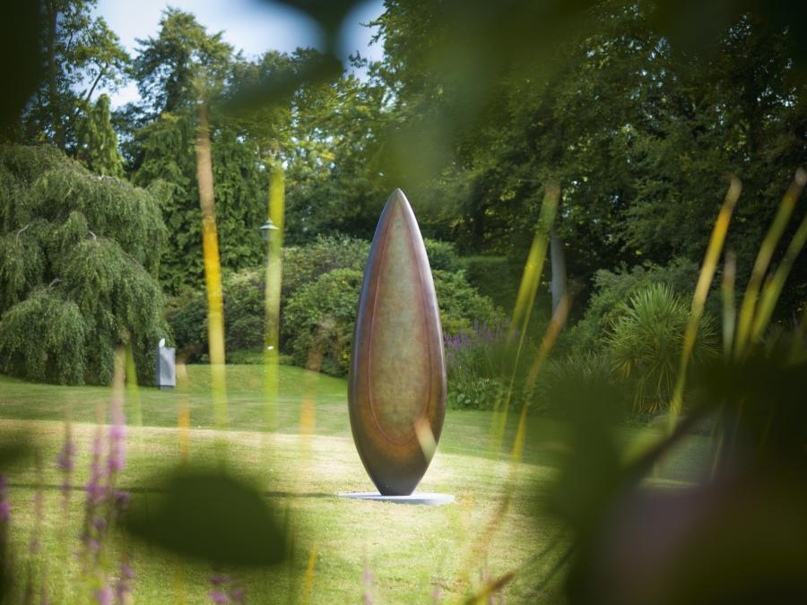 Monumental bronze sculpture by Dominic Welch, Rising Form VI, standing over 2.5 metres tall. The elongated leaf-like form features a smooth, tapering silhouette with a central turquoise-green patinated core against rich bronze tones. A powerful outdoor or architectural piece, it radiates elegance, presence, and organic energy.