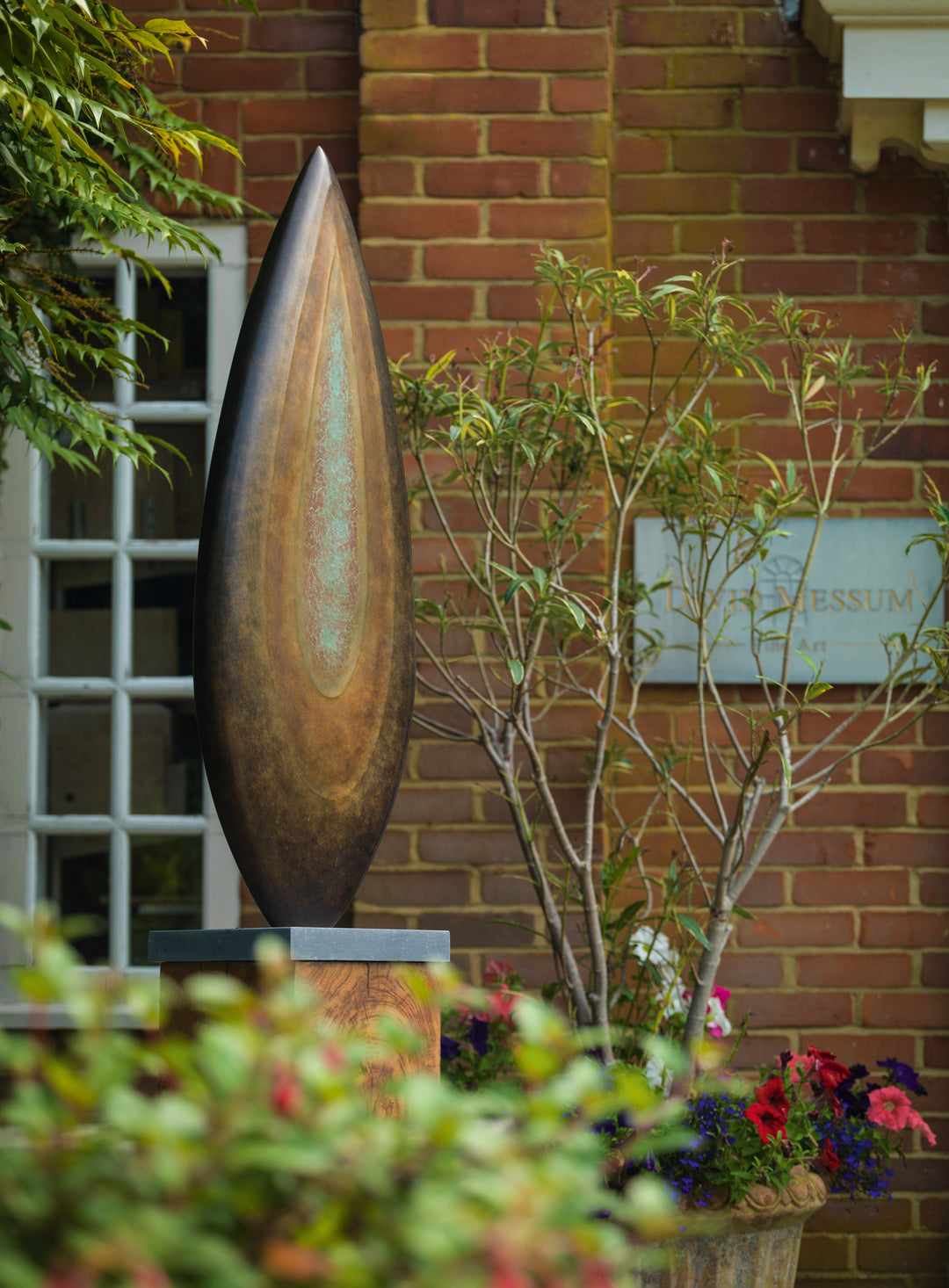 Abstract bronze sculpture Rising Form X by Dominic Welch, shaped like an elongated seed pod with smooth organic contours. The surface patina shifts from warm bronze to subtle green tones, mounted on a natural wood plinth, set within a garden courtyard.