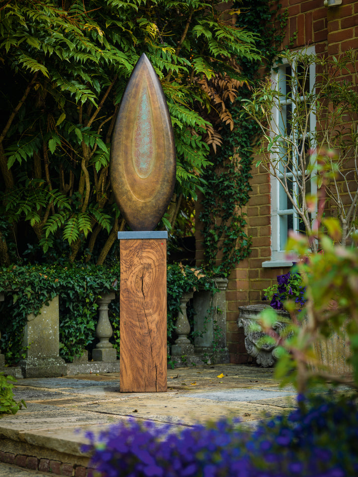 Abstract bronze sculpture Rising Form X by Dominic Welch, shaped like an elongated seed pod with smooth organic contours. The surface patina shifts from warm bronze to subtle green tones, mounted on a natural wood plinth, set within a garden courtyard.