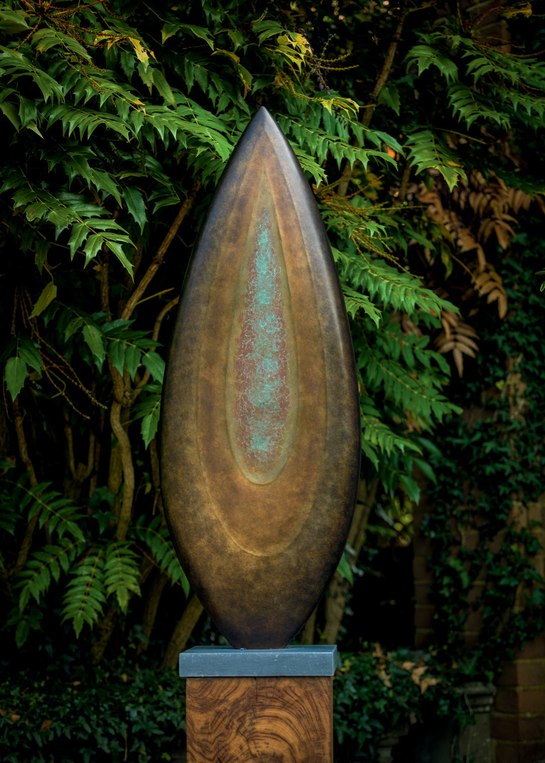 Abstract bronze sculpture Rising Form X by Dominic Welch, shaped like an elongated seed pod with smooth organic contours. The surface patina shifts from warm bronze to subtle green tones, mounted on a natural wood plinth, set within a garden courtyard.