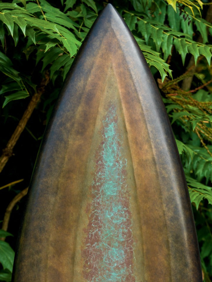 Abstract bronze sculpture Rising Form X by Dominic Welch, shaped like an elongated seed pod with smooth organic contours. The surface patina shifts from warm bronze to subtle green tones, mounted on a natural wood plinth, set within a garden courtyard.