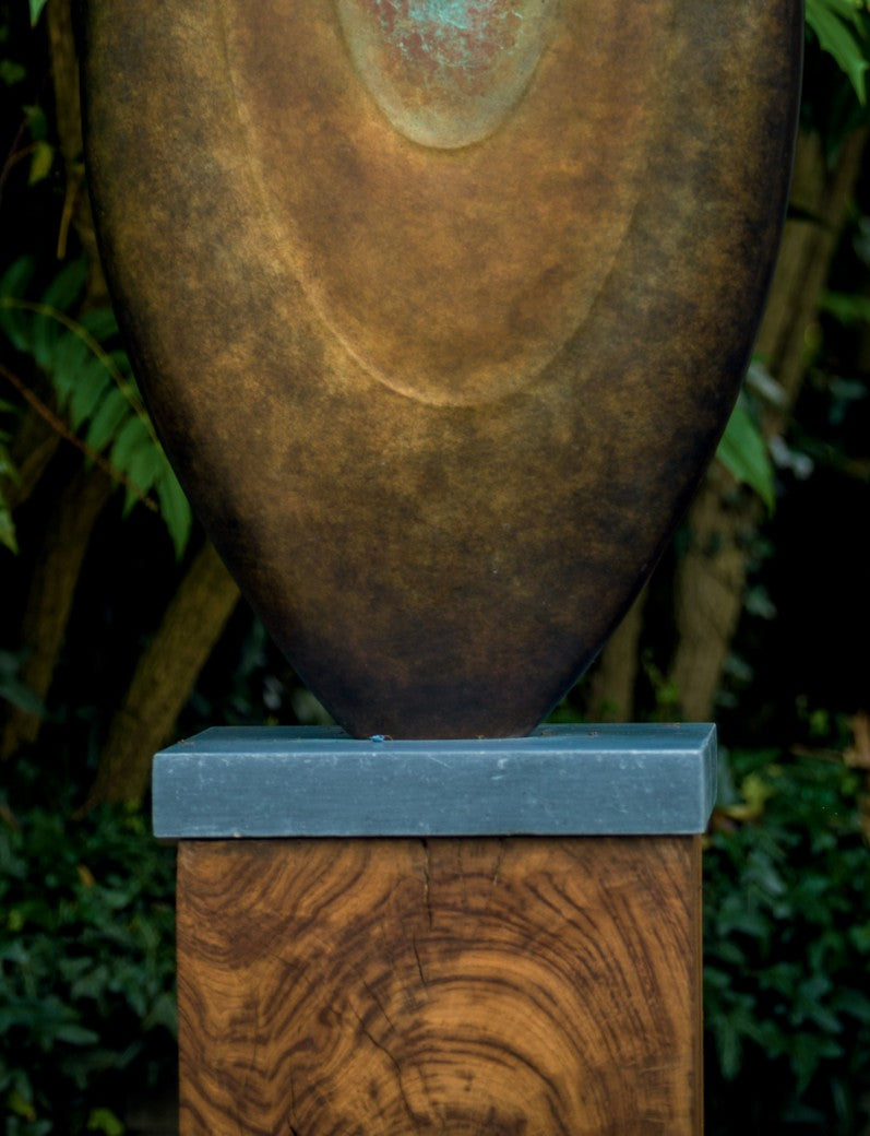 Abstract bronze sculpture Rising Form X by Dominic Welch, shaped like an elongated seed pod with smooth organic contours. The surface patina shifts from warm bronze to subtle green tones, mounted on a natural wood plinth, set within a garden courtyard.