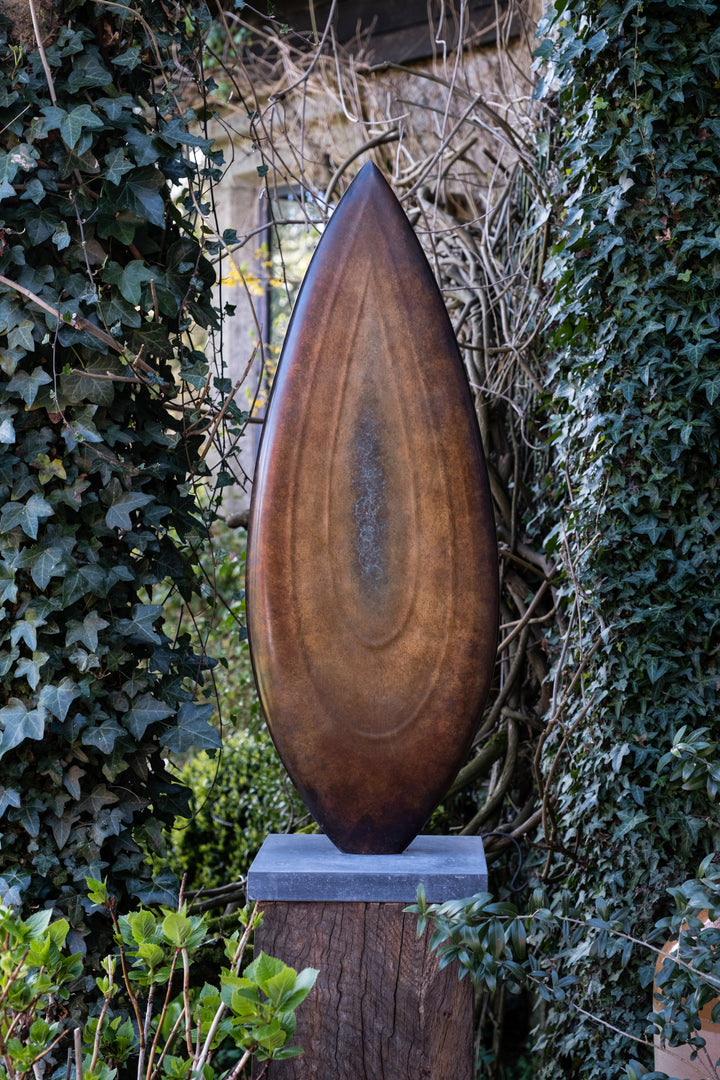 Abstract bronze sculpture Rising Form X by Dominic Welch, shaped like an elongated seed pod with smooth organic contours. The surface patina shifts from warm bronze to subtle green tones, mounted on a natural wood plinth, set within a garden courtyard.