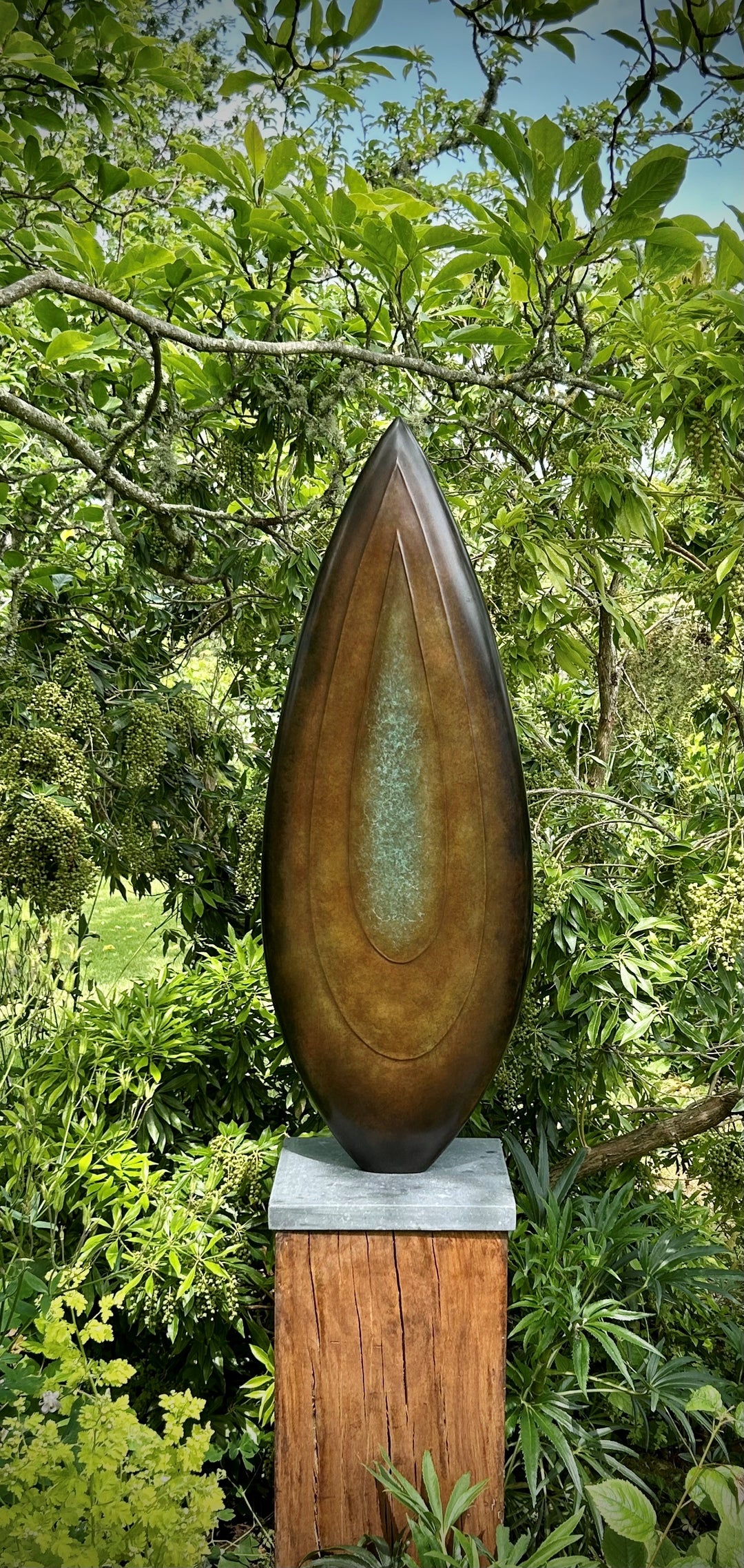 Abstract bronze sculpture Rising Form X by Dominic Welch, shaped like an elongated seed pod with smooth organic contours. The surface patina shifts from warm bronze to subtle green tones, mounted on a natural wood plinth, set within a garden courtyard.