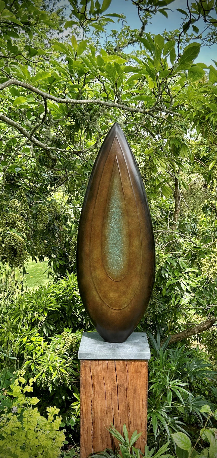 Abstract bronze sculpture Rising Form X by Dominic Welch, shaped like an elongated seed pod with smooth organic contours. The surface patina shifts from warm bronze to subtle green tones, mounted on a natural wood plinth, set within a garden courtyard.