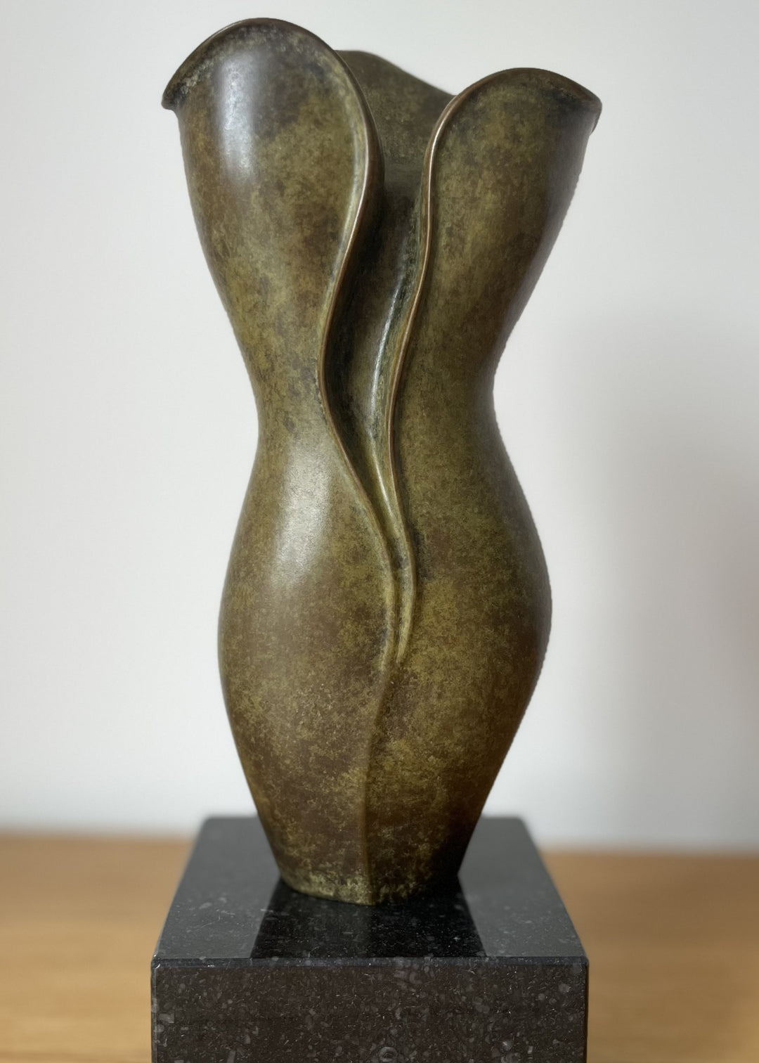 Elegant small bronze sculpture with a fluid, petal-like form and gently folding curves, finished in an earthy green-brown patina with subtle mottling, mounted on a polished black stone base.