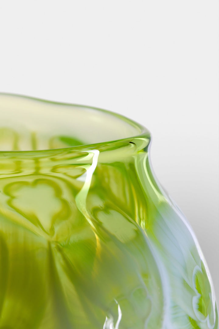 Green hand-blown glass vessel featuring a textured surface with embossed floral motifs and a gently pinched base, shown against a white reflective background.