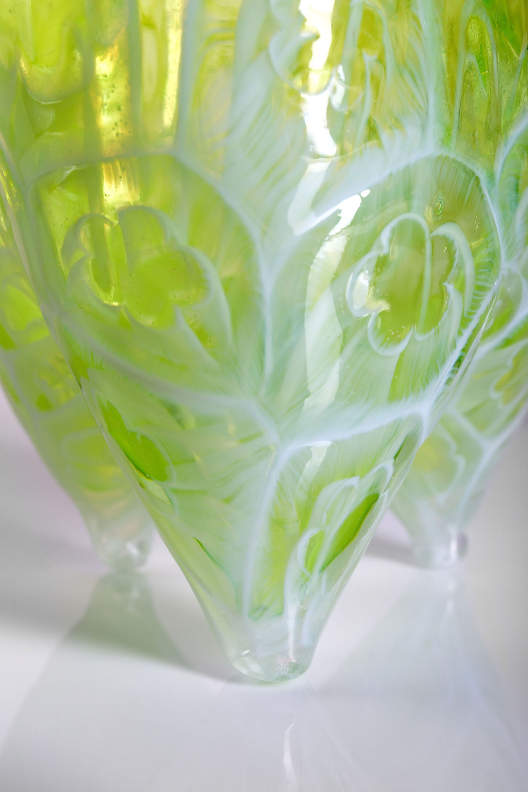 Tall handblown green glass sculpture with embossed floral motifs and a softly asymmetrical, pod-like form, resting on a reflective surface.