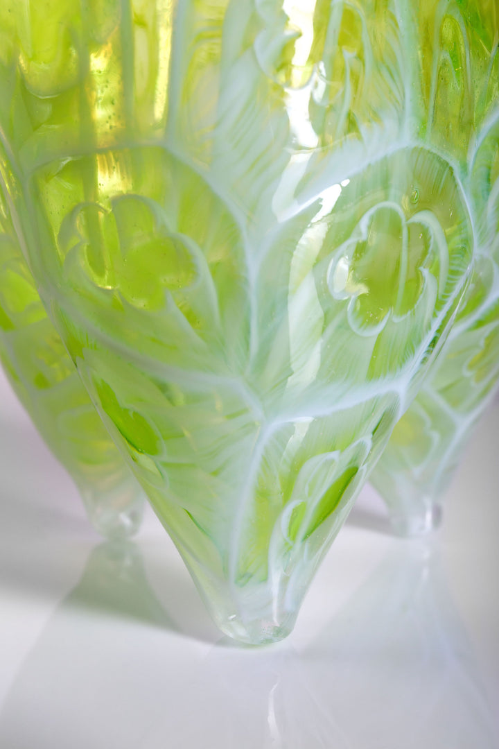 Tall handblown green glass sculpture with embossed floral motifs and a softly asymmetrical, pod-like form, resting on a reflective surface.