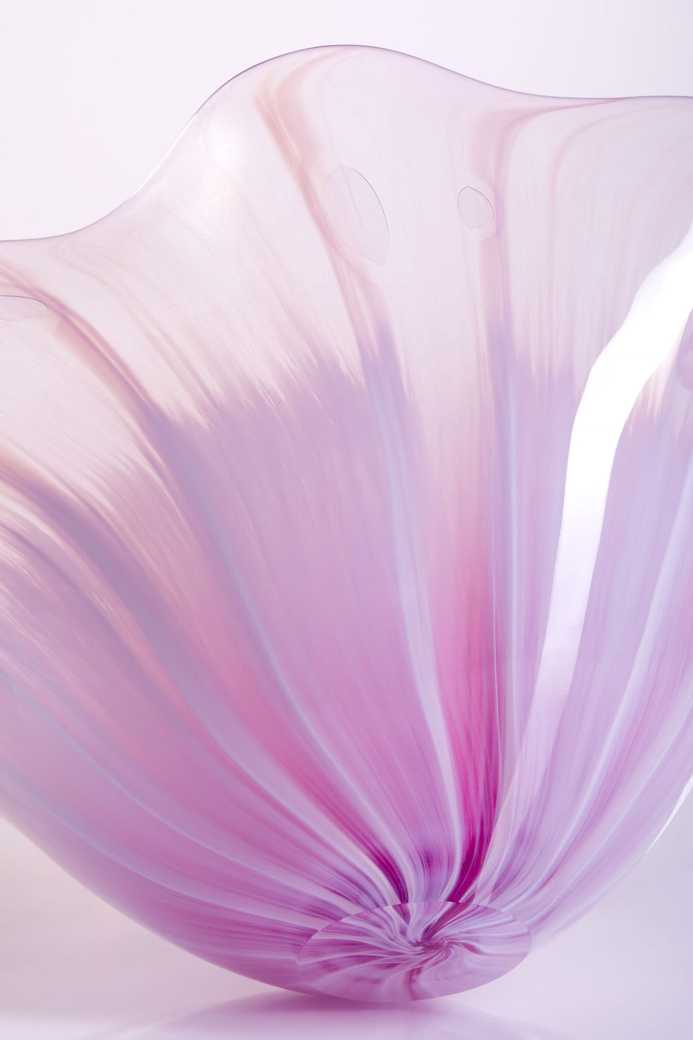 Delicate, translucent handblown glass vessel in soft pink and lavender hues, fanned open like a flower, set against a light background with a mirrored reflection.