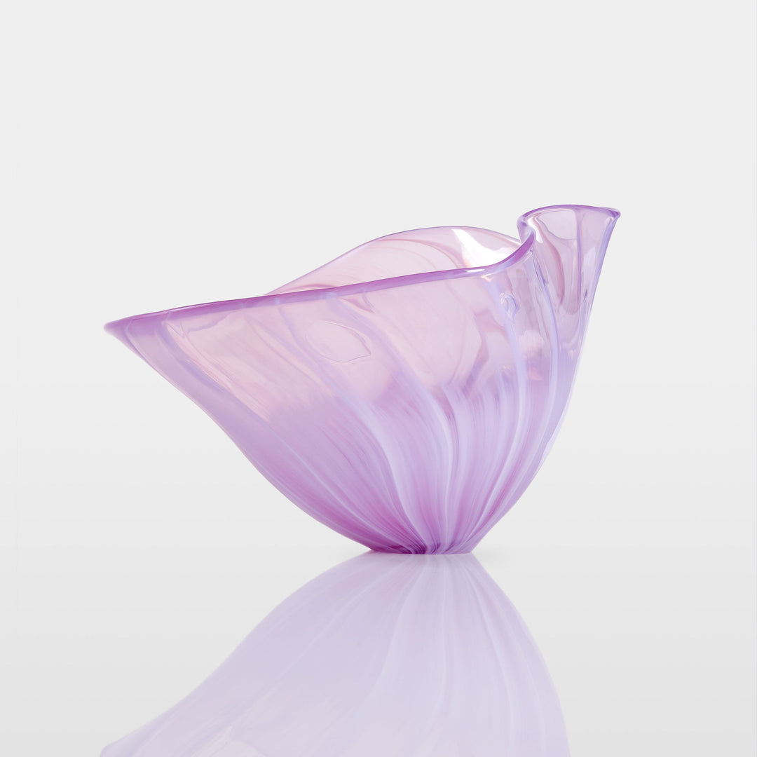 Delicate, translucent handblown glass vessel in soft pink and lavender hues, fanned open like a flower, set against a light background with a mirrored reflection.