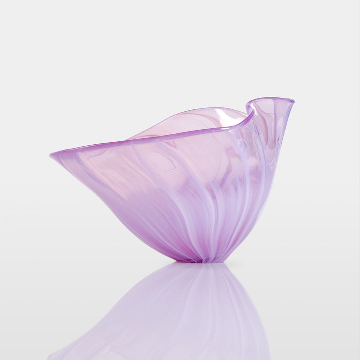 Delicate, translucent handblown glass vessel in soft pink and lavender hues, fanned open like a flower, set against a light background with a mirrored reflection.