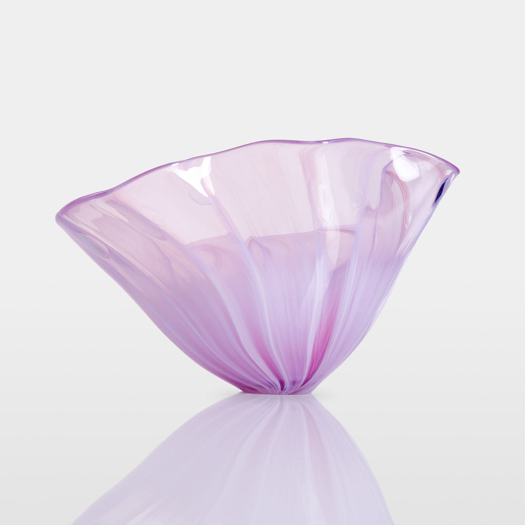 Delicate, translucent handblown glass vessel in soft pink and lavender hues, fanned open like a flower, set against a light background with a mirrored reflection.