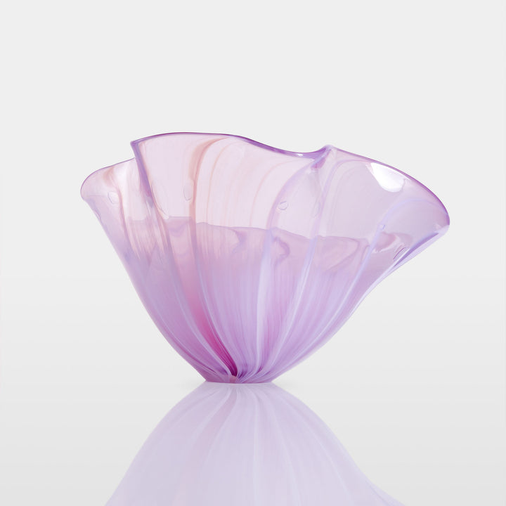 Delicate, translucent handblown glass vessel in soft pink and lavender hues, fanned open like a flower, set against a light background with a mirrored reflection.