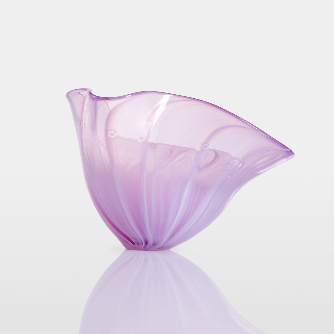Delicate, translucent handblown glass vessel in soft pink and lavender hues, fanned open like a flower, set against a light background with a mirrored reflection.
