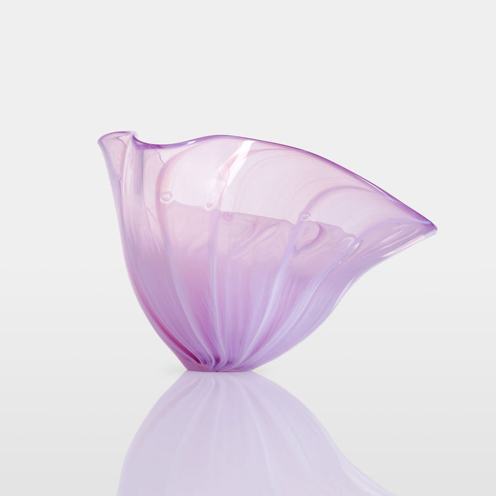 Delicate, translucent handblown glass vessel in soft pink and lavender hues, fanned open like a flower, set against a light background with a mirrored reflection.