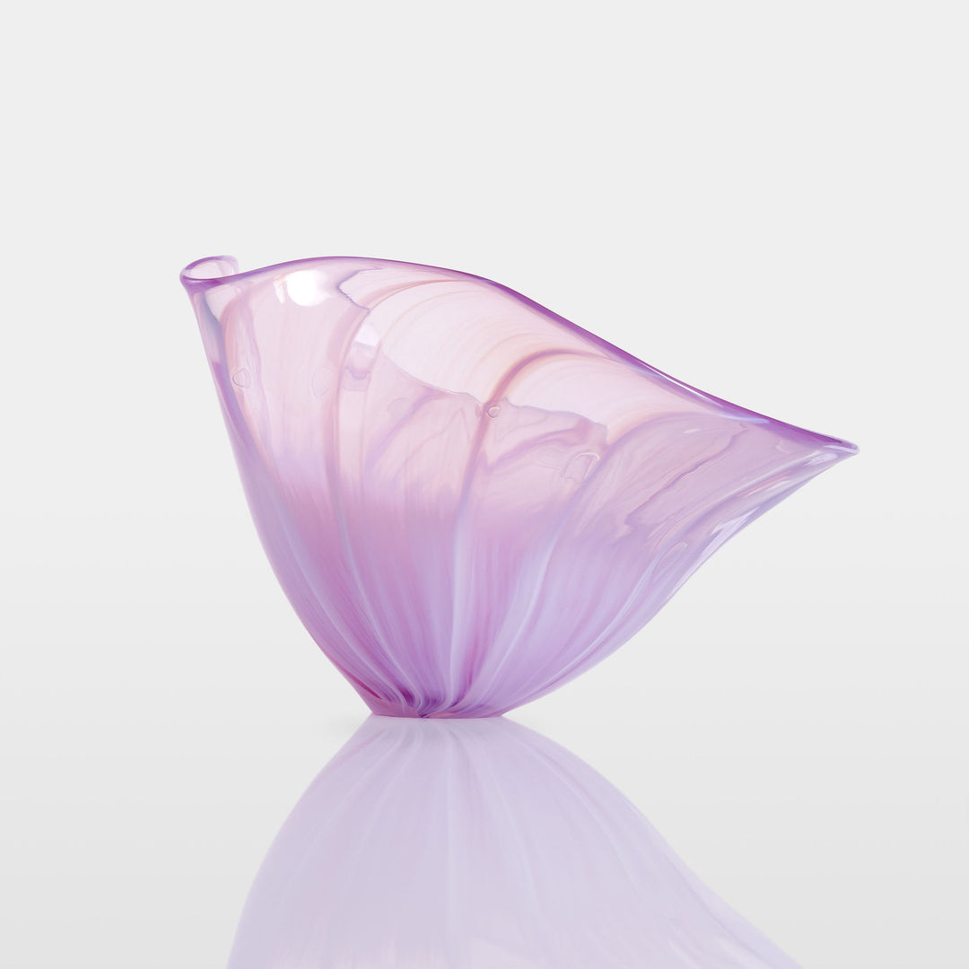 Delicate, translucent handblown glass vessel in soft pink and lavender hues, fanned open like a flower, set against a light background with a mirrored reflection.