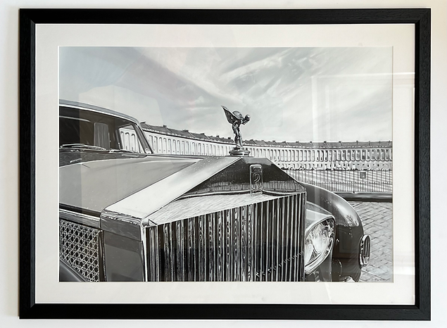 Monochrome pastel artwork depicting the front grille and bonnet of a Rolls-Royce with the Spirit of Ecstasy ornament in sharp focus, positioned against the sweeping curve of Bath’s Royal Crescent, with polished chrome, sculpted metal and architectural stonework rendered through subtle tonal shifts, light and shadow.
