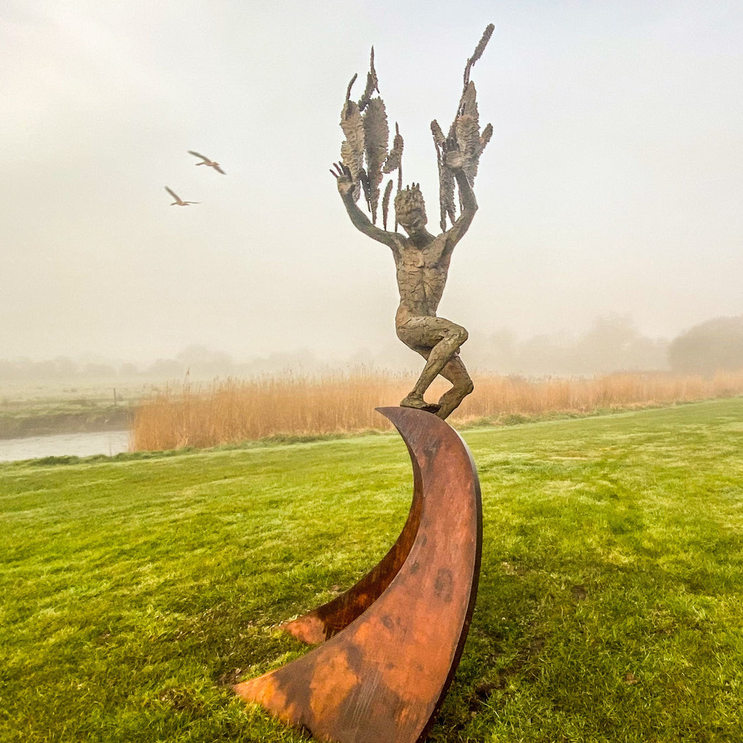 Monumental outdoor bronze sculpture by Nicola Godden — a dramatic, winged male figure with raised arms, climbing and balancing on a sweeping rusted steel crescent. The figure’s body has a textured verdigris green and earthy bronze patina with highlights of gold and brown, contrasting with the deep weathered red-orange steel base. Photographed in an open grassy field with blue sky.