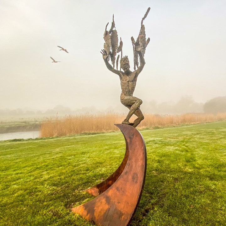 Monumental outdoor bronze sculpture by Nicola Godden — a dramatic, winged male figure with raised arms, climbing and balancing on a sweeping rusted steel crescent. The figure’s body has a textured verdigris green and earthy bronze patina with highlights of gold and brown, contrasting with the deep weathered red-orange steel base. Photographed in an open grassy field with blue sky.