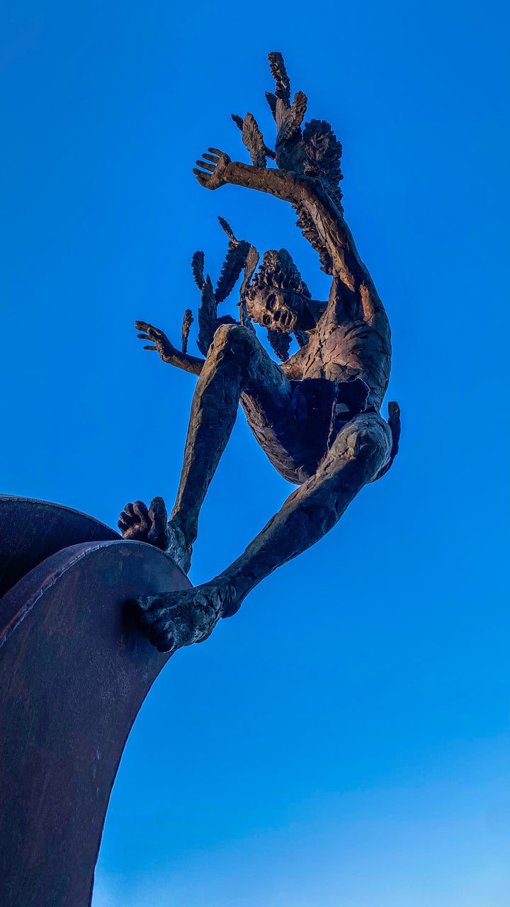 Monumental outdoor bronze sculpture by Nicola Godden — a dramatic, winged male figure with raised arms, climbing and balancing on a sweeping rusted steel crescent. The figure’s body has a textured verdigris green and earthy bronze patina with highlights of gold and brown, contrasting with the deep weathered red-orange steel base. Photographed in an open grassy field with blue sky.