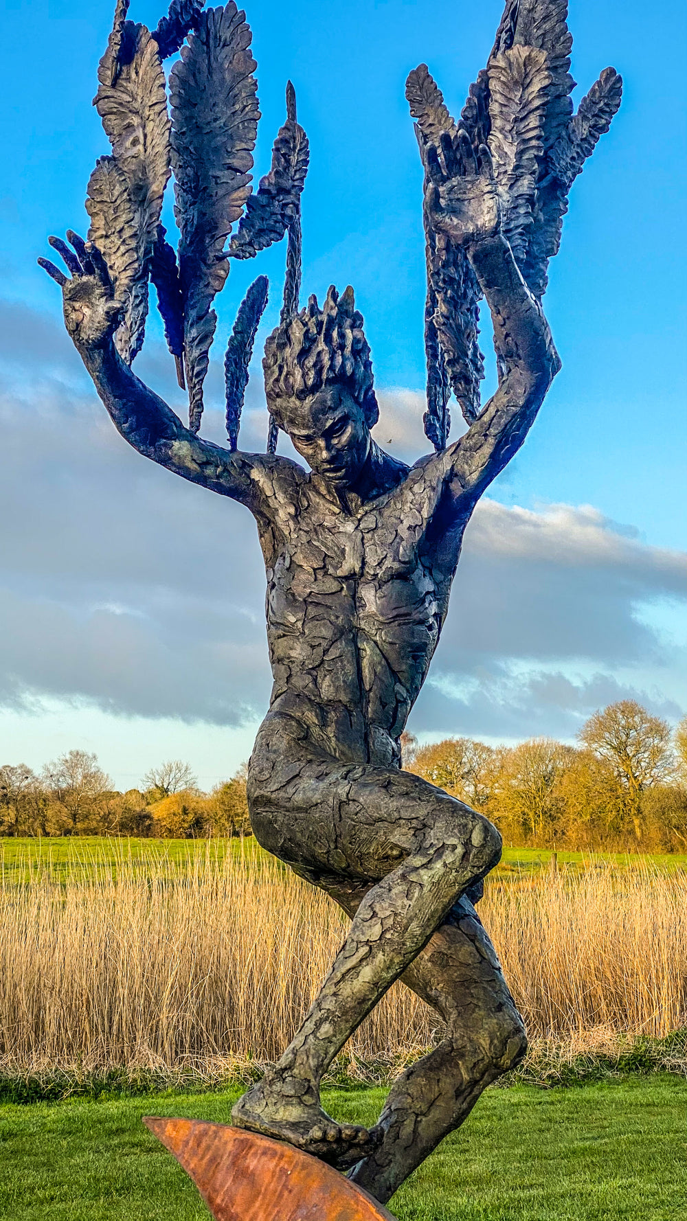 Monumental outdoor bronze sculpture by Nicola Godden — a dramatic, winged male figure with raised arms, climbing and balancing on a sweeping rusted steel crescent. The figure’s body has a textured verdigris green and earthy bronze patina with highlights of gold and brown, contrasting with the deep weathered red-orange steel base. Photographed in an open grassy field with blue sky.