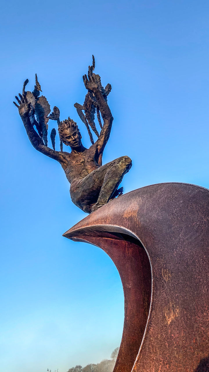Monumental outdoor bronze sculpture by Nicola Godden — a dramatic, winged male figure with raised arms, climbing and balancing on a sweeping rusted steel crescent. The figure’s body has a textured verdigris green and earthy bronze patina with highlights of gold and brown, contrasting with the deep weathered red-orange steel base. Photographed in an open grassy field with blue sky.