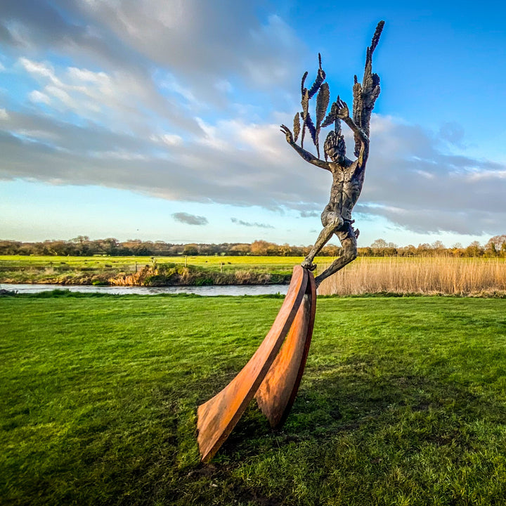 Monumental outdoor bronze sculpture by Nicola Godden — a dramatic, winged male figure with raised arms, climbing and balancing on a sweeping rusted steel crescent. The figure’s body has a textured verdigris green and earthy bronze patina with highlights of gold and brown, contrasting with the deep weathered red-orange steel base. Photographed in an open grassy field with blue sky.