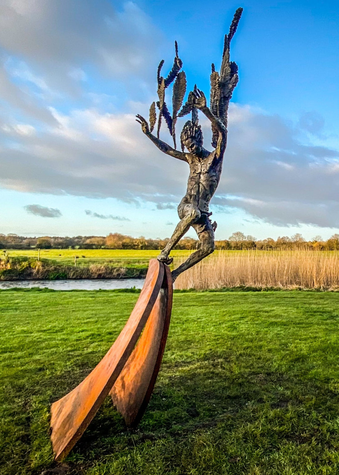 Monumental outdoor bronze sculpture by Nicola Godden — a dramatic, winged male figure with raised arms, climbing and balancing on a sweeping rusted steel crescent. The figure’s body has a textured verdigris green and earthy bronze patina with highlights of gold and brown, contrasting with the deep weathered red-orange steel base. Photographed in an open grassy field with blue sky.