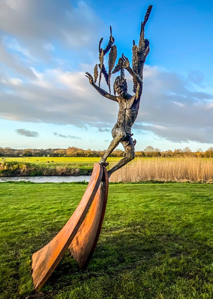 Monumental outdoor bronze sculpture by Nicola Godden — a dramatic, winged male figure with raised arms, climbing and balancing on a sweeping rusted steel crescent. The figure’s body has a textured verdigris green and earthy bronze patina with highlights of gold and brown, contrasting with the deep weathered red-orange steel base. Photographed in an open grassy field with blue sky.