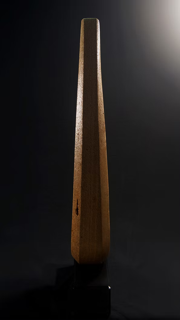 Tall Purbeck stone sculpture with a central vertical opening and tapered peak, standing on a black granite base, showing fossil textures and carved geometry against a dark background.