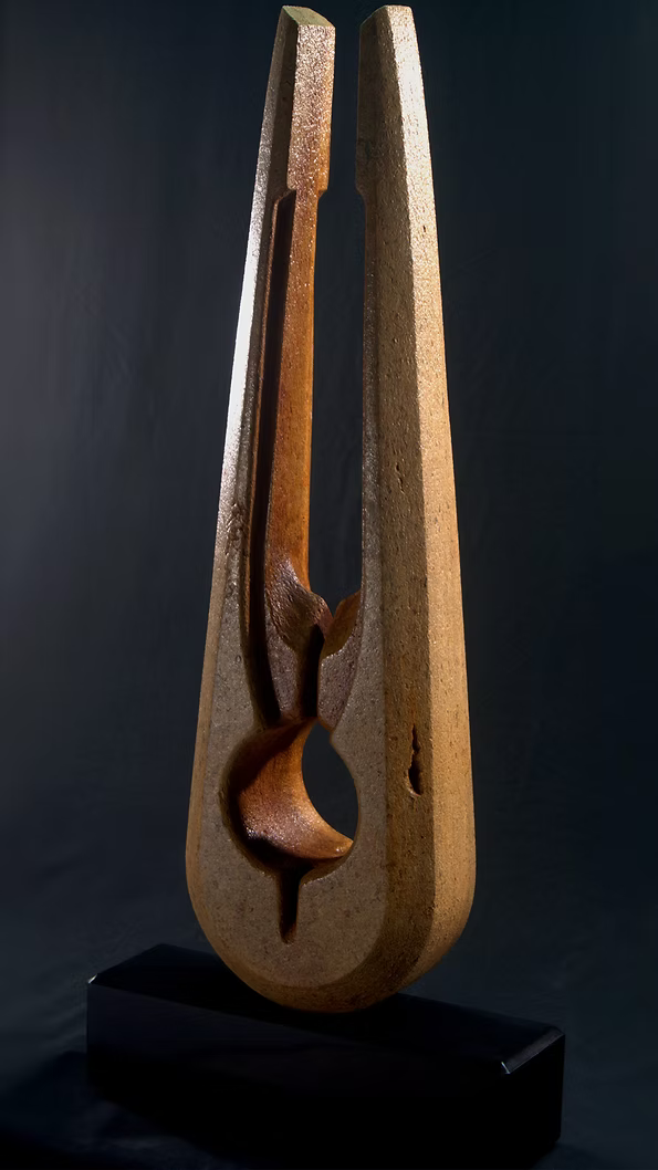 Tall Purbeck stone sculpture with a central vertical opening and tapered peak, standing on a black granite base, showing fossil textures and carved geometry against a dark background.