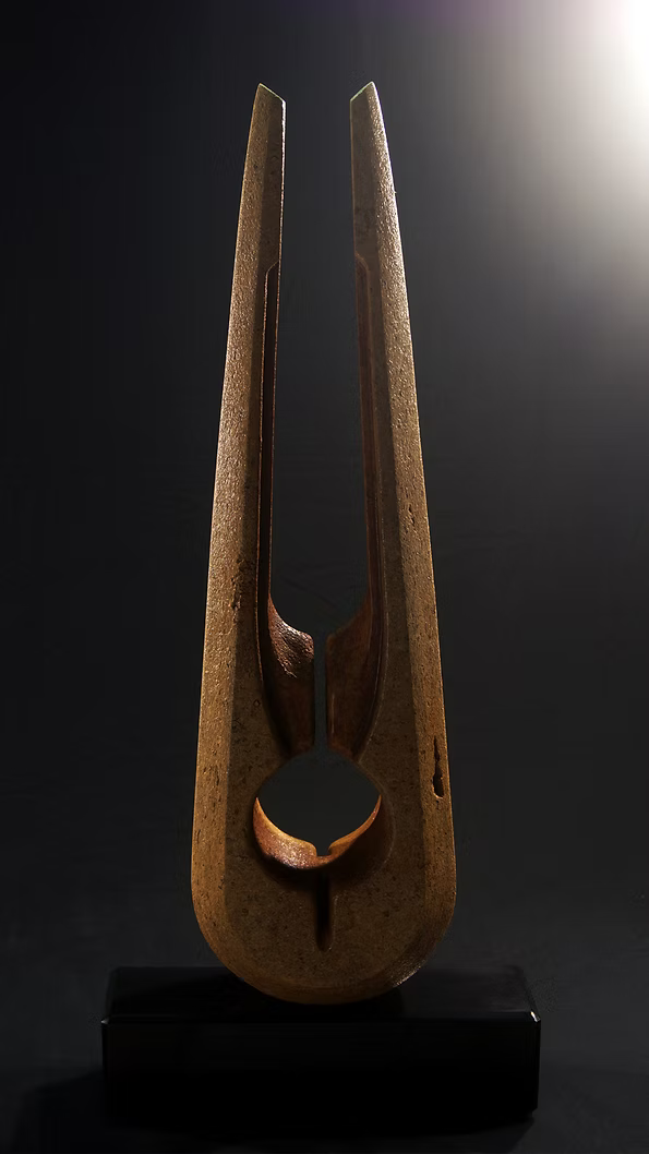 Tall Purbeck stone sculpture with a central vertical opening and tapered peak, standing on a black granite base, showing fossil textures and carved geometry against a dark background.