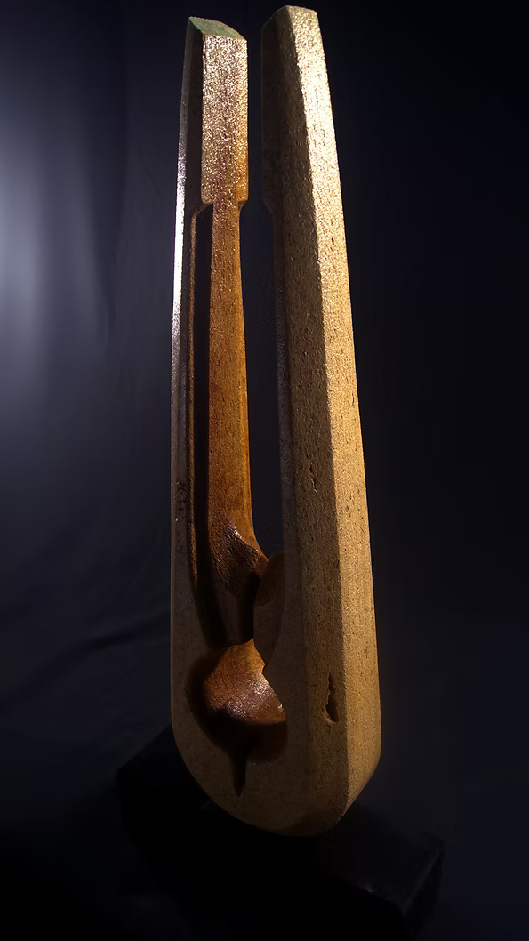 Tall Purbeck stone sculpture with a central vertical opening and tapered peak, standing on a black granite base, showing fossil textures and carved geometry against a dark background.