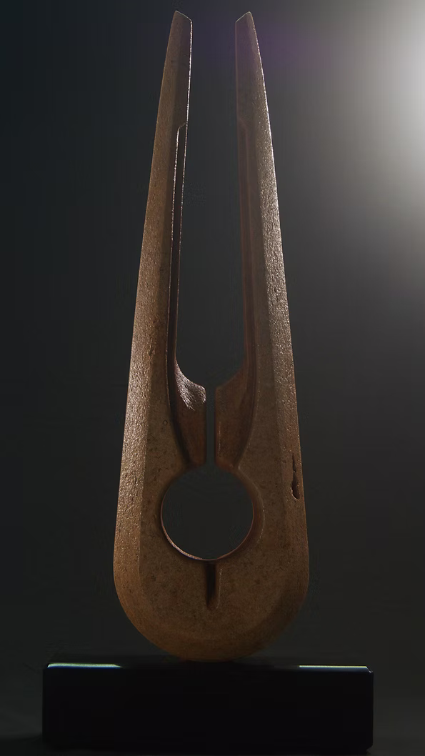 Tall Purbeck stone sculpture with a central vertical opening and tapered peak, standing on a black granite base, showing fossil textures and carved geometry against a dark background.