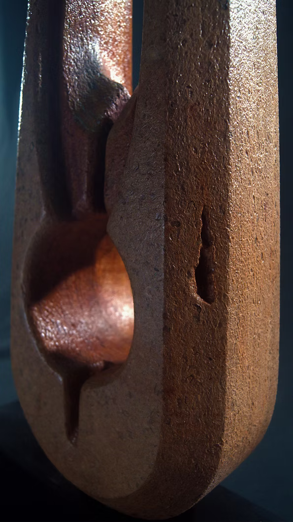 Tall Purbeck stone sculpture with a central vertical opening and tapered peak, standing on a black granite base, showing fossil textures and carved geometry against a dark background.