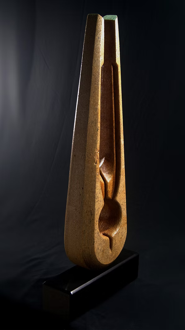 Tall Purbeck stone sculpture with a central vertical opening and tapered peak, standing on a black granite base, showing fossil textures and carved geometry against a dark background.