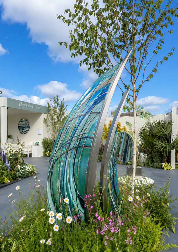 Carrie-Anne Funnell glass and stainless steel sculpture Sail Away, featuring two large curved sail-like forms with layered blue, turquoise and aqua glass framed in polished stainless steel, displayed within a landscaped outdoor garden setting.