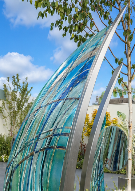 Carrie-Anne Funnell glass and stainless steel sculpture Sail Away, featuring two large curved sail-like forms with layered blue, turquoise and aqua glass framed in polished stainless steel, displayed within a landscaped outdoor garden setting.