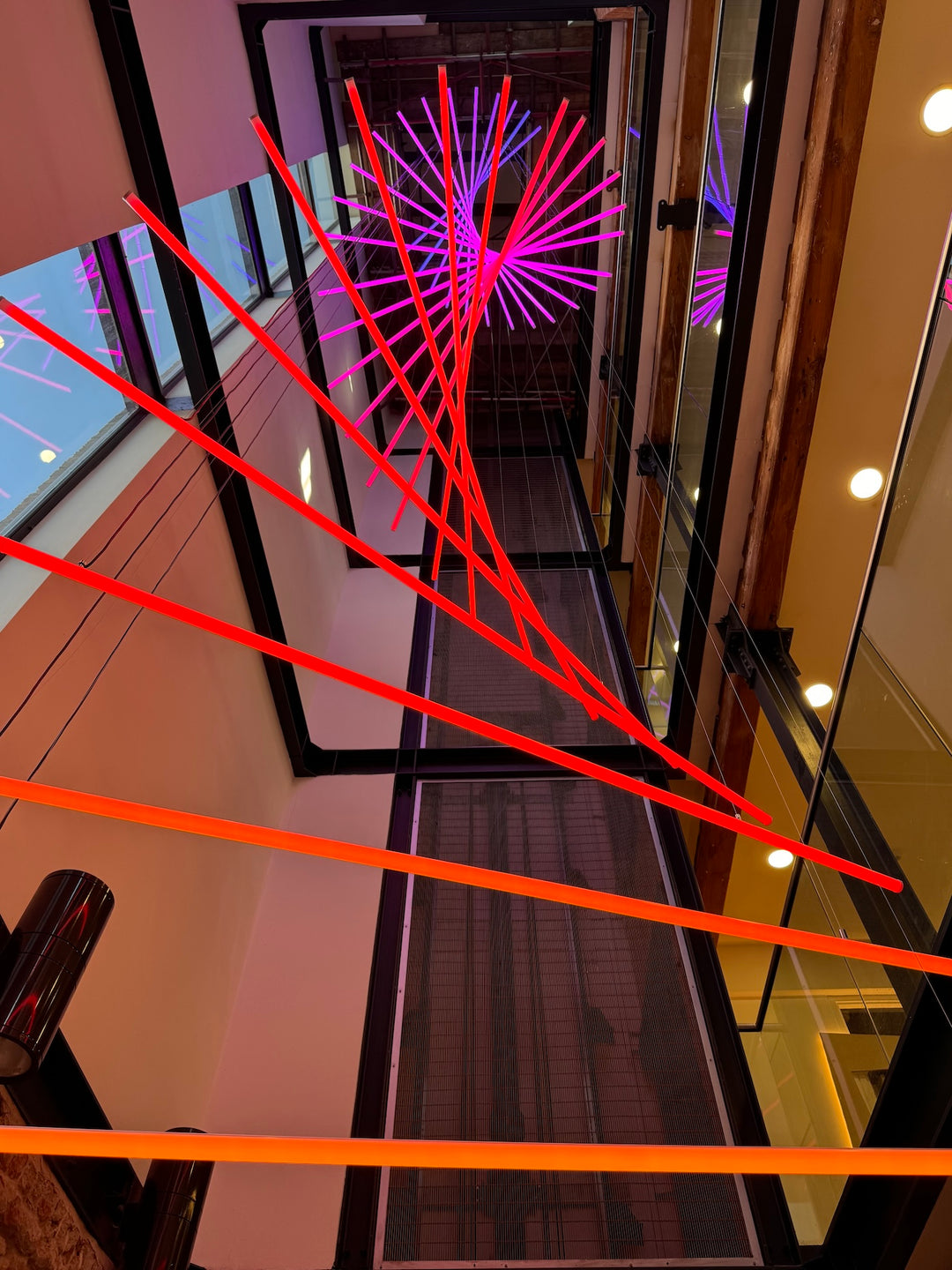Suspended spiral light installation composed of intersecting illuminated linear elements arranged around a vertical stairwell void, with coloured light bars radiating and twisting through space to echo the geometry and movement of the surrounding architecture, creating layered reflections and depth within the entrance atrium.
