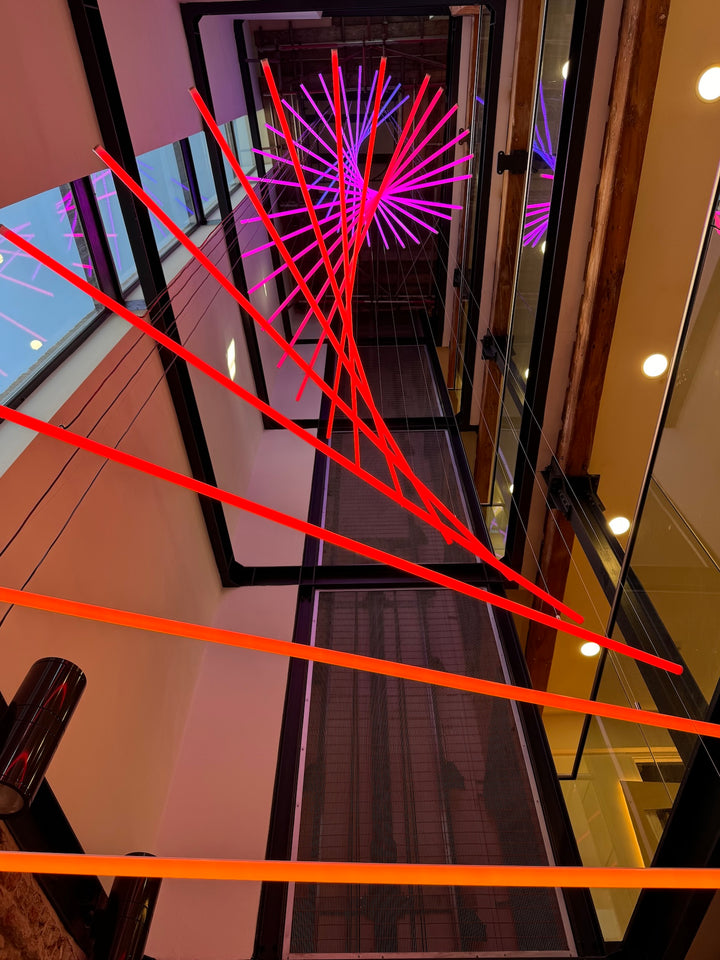 Suspended spiral light installation composed of intersecting illuminated linear elements arranged around a vertical stairwell void, with coloured light bars radiating and twisting through space to echo the geometry and movement of the surrounding architecture, creating layered reflections and depth within the entrance atrium.