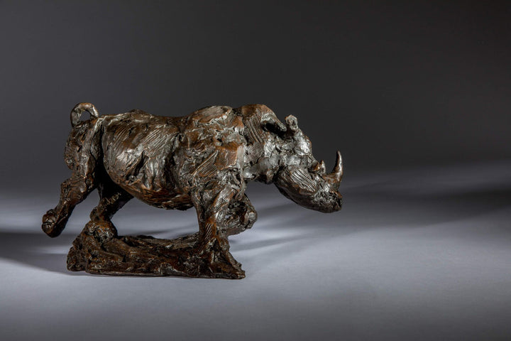 Bronze sculpture of a white rhino mid-charge, captured with bold textures and muscular form, evoking movement and strength.