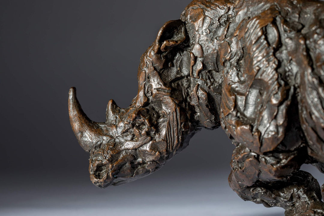 Bronze sculpture of a white rhino mid-charge, captured with bold textures and muscular form, evoking movement and strength.